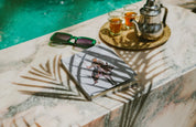 Moroccan tea set, ethnik illustrated notebook, and sunglasses by a pool on a marble surface.