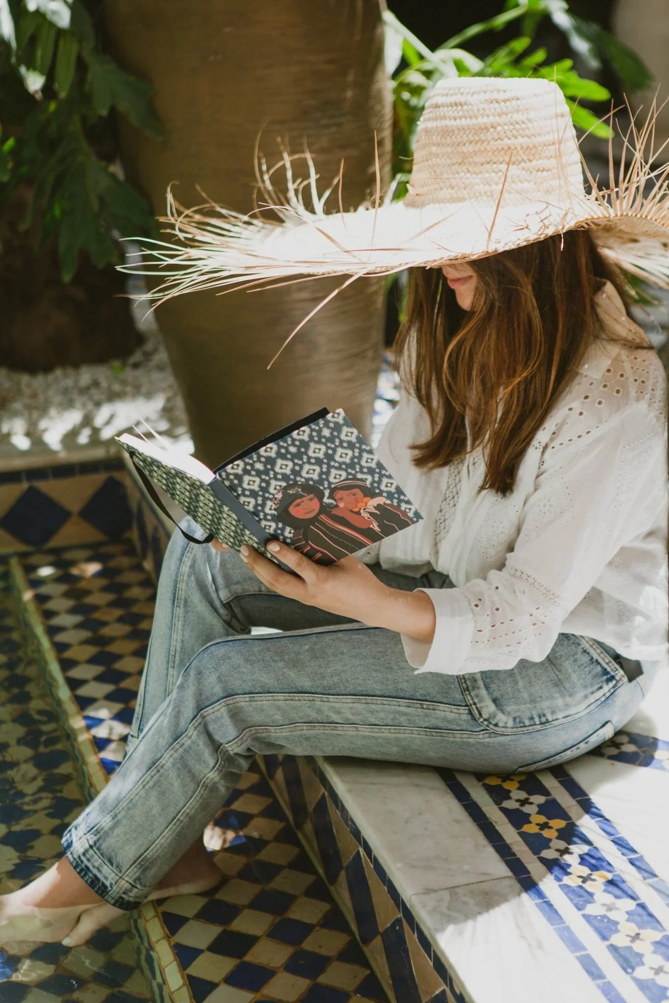 Woman in straw hat reads ethnik Moroccan illustrated notebook by mosaic pool, natural light