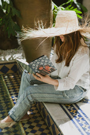 Woman in straw hat reads ethnik Moroccan illustrated notebook by mosaic pool, natural light