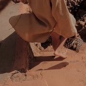 Person in beige robe walking barefoot on stone stairs, holding Ethnik illustrated pouch, Morocco