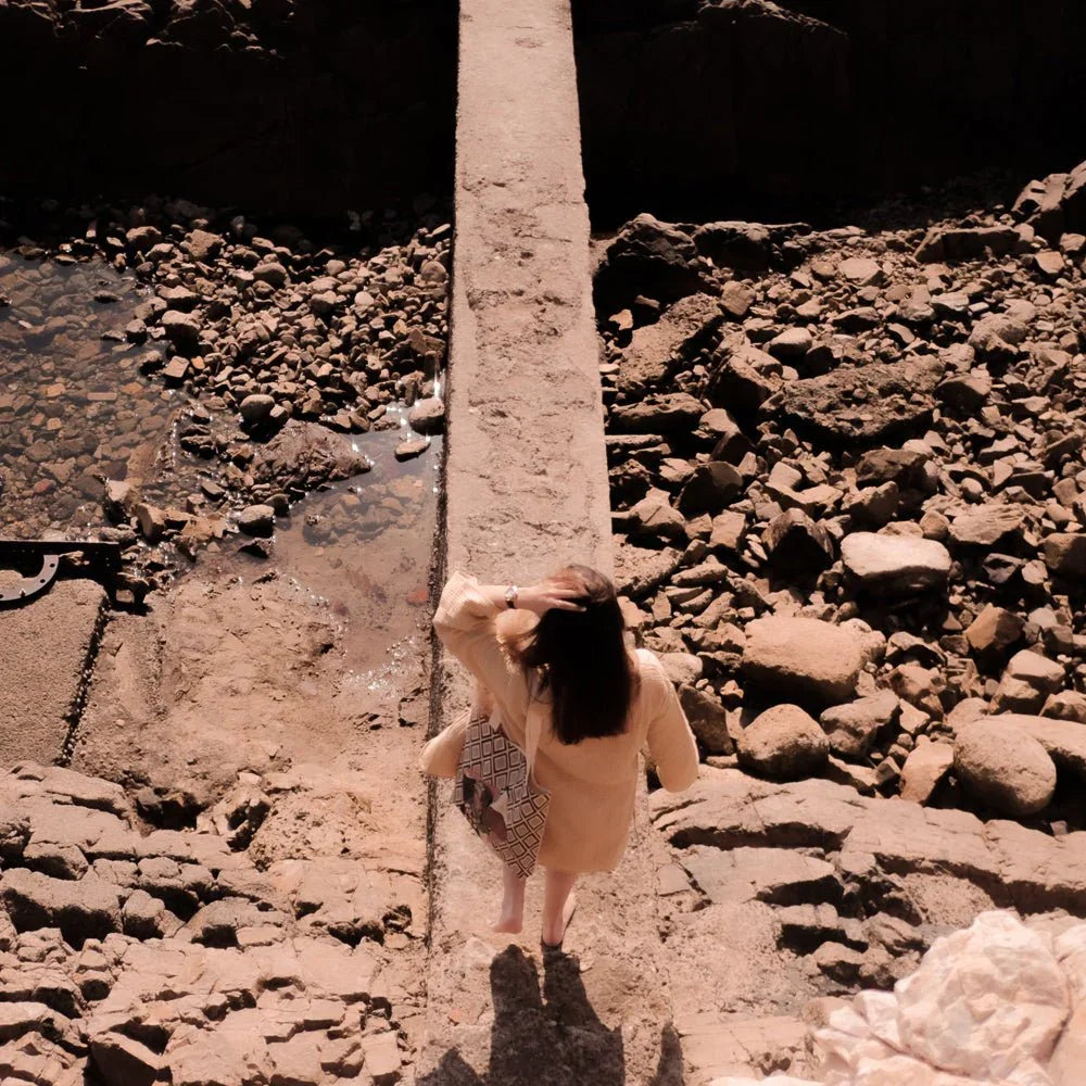 Woman in beige coat on rocky path holding patterned ethnik tote bag, natural sunlight