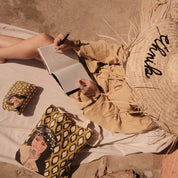 Woman sketching in notebook outdoors, wearing ethnik straw hat, with Moroccan illustrated tote and pouch.